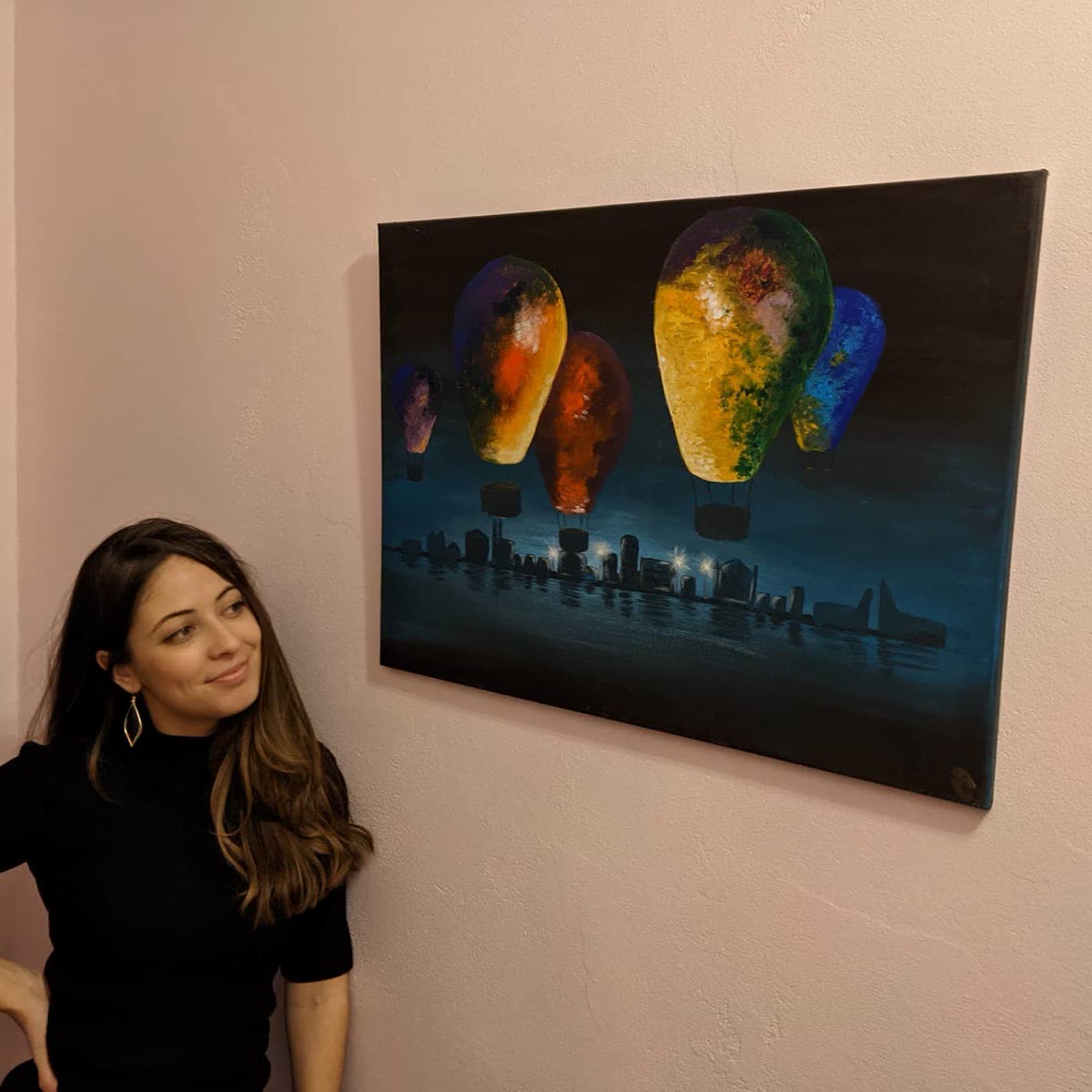 A dark canvas of hot-air balloons rising over a city skyline, hanging on a wall with the artist beside it.